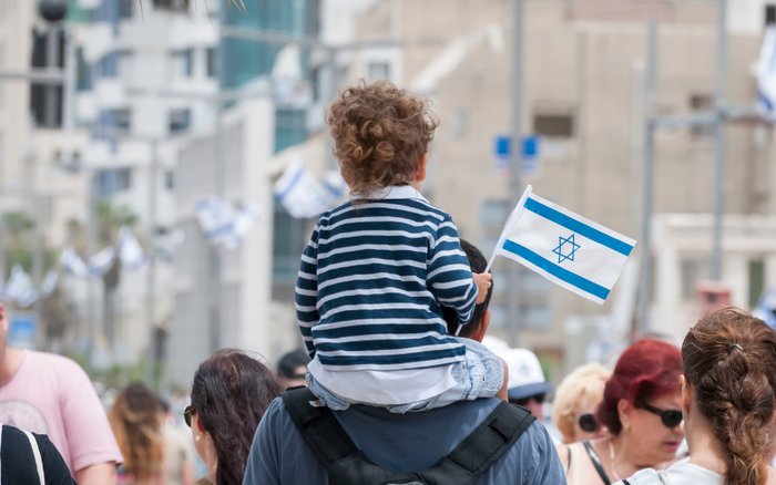 Child on an adult's shoulders holding an Israeli flag in a crowd at an Independence Day celebration